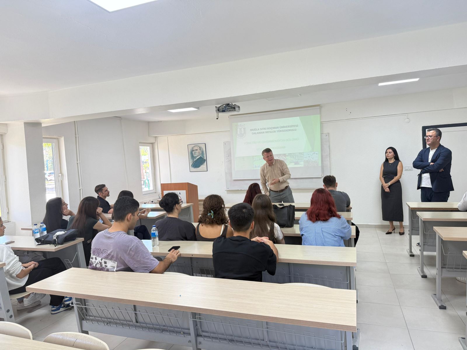 Muğla Sıtkı Koçman Üniversitesi Dalaman Meslek Yüksek Okulunda derslikte lojistik programı tanıtımı; bir konuşmacı anlatıyor, iki kişi ayakta, bunlar eğitimcilerdir, öğrenciler dinliyor.